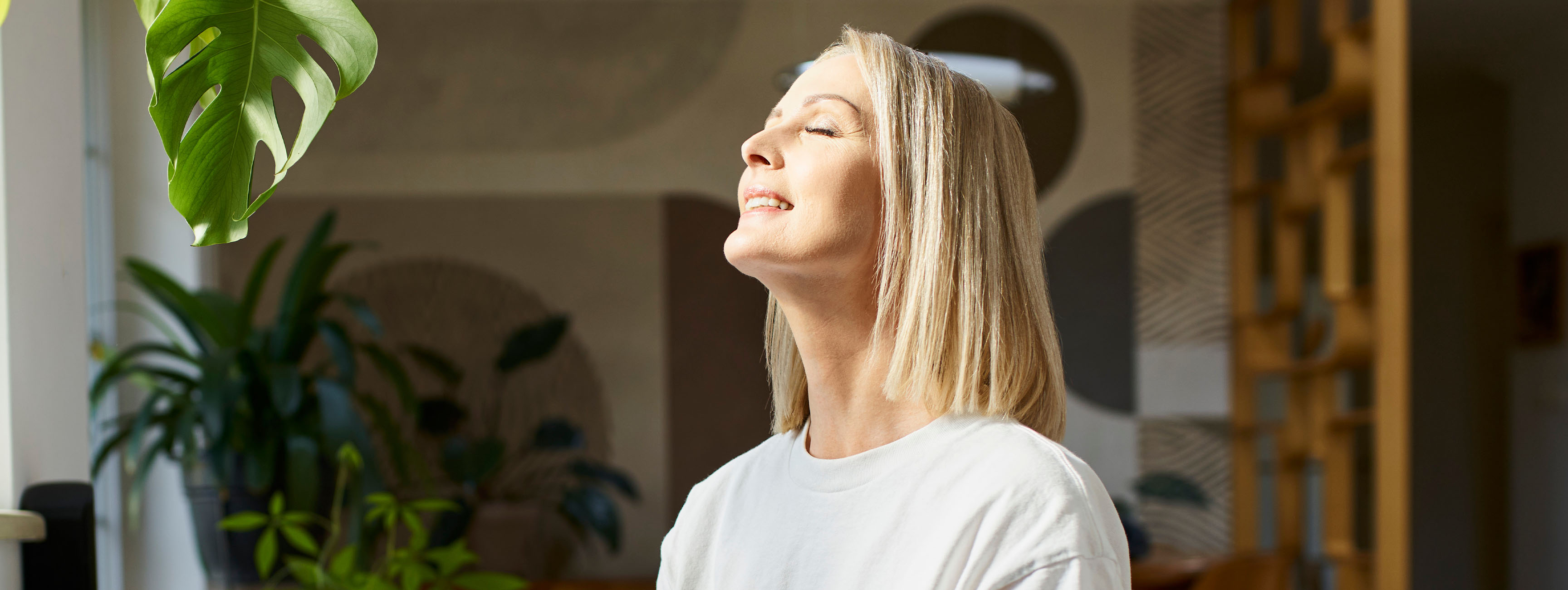 A smiling woman in her living room faces a bright window with her eyes closed
