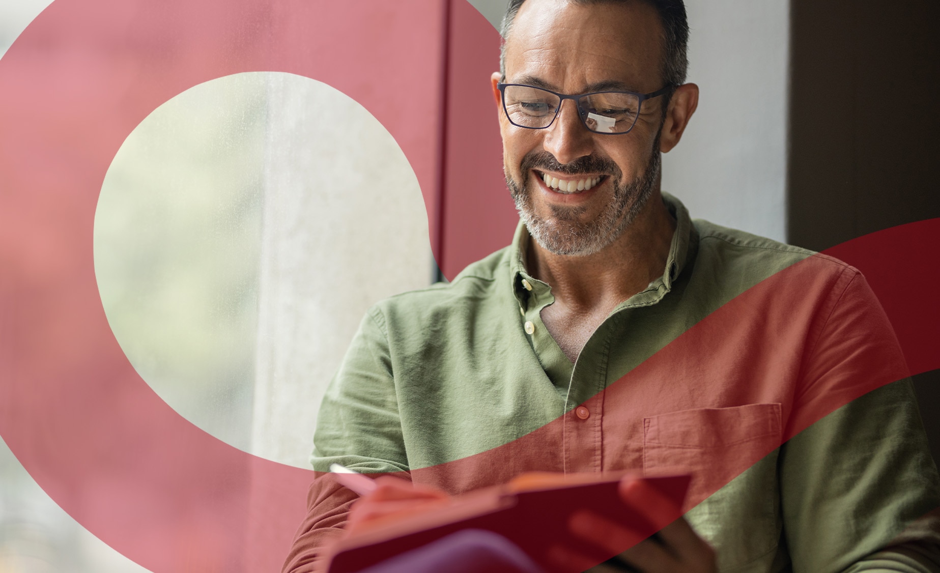 A smiling man sits by a window writing in a paper journal.