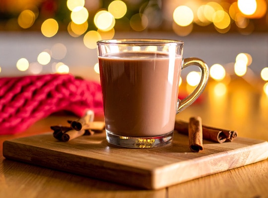 Glass mug of steaming Gingerbread Hot Chocolate made with KitKat, served on a wooden board with cinnamon sticks, festive ligh