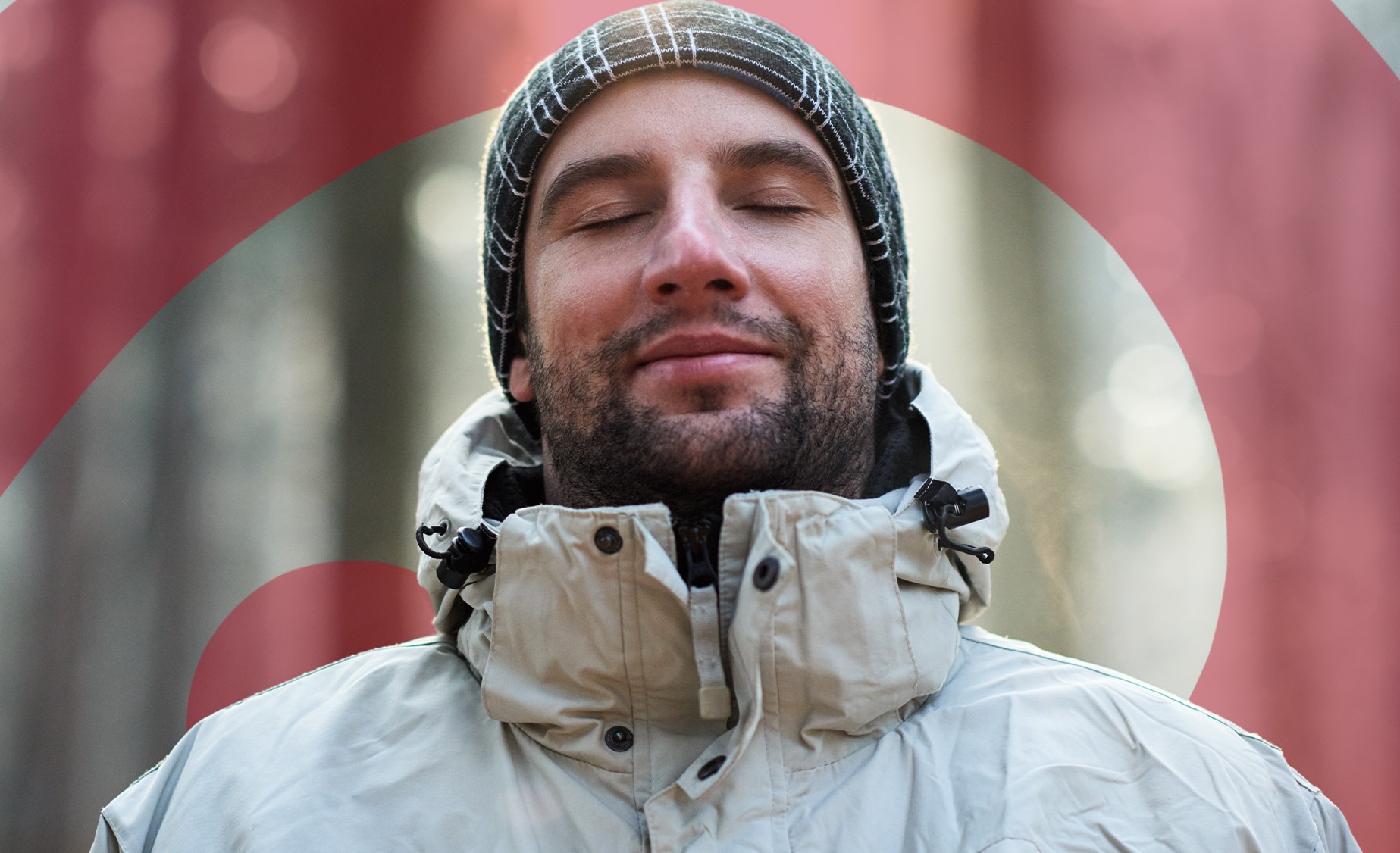 A smiling man with his eyes closed standing outside in the woods wearing a beanie hat  