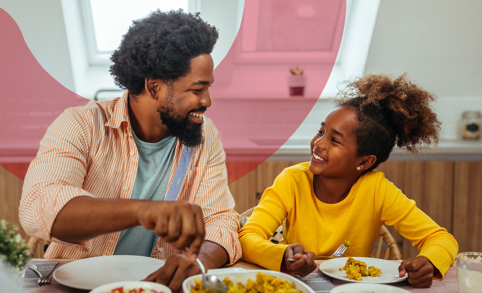 A father laughing with his daughter over lunch indoors at a wooden table with windows in the background 