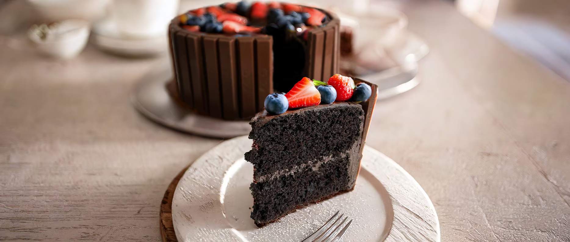 A dark chocolate cake topped with fruit and edged with a ring of KitKats, with the  whole cake in the background and a slice.