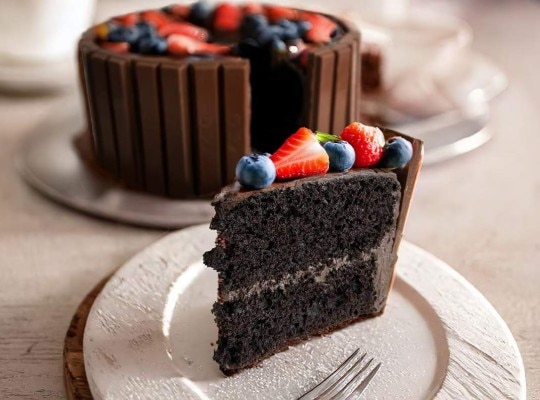 A dark chocolate cake topped with fruit and edged with a ring of KitKats, with the  whole cake in the background and a slice.