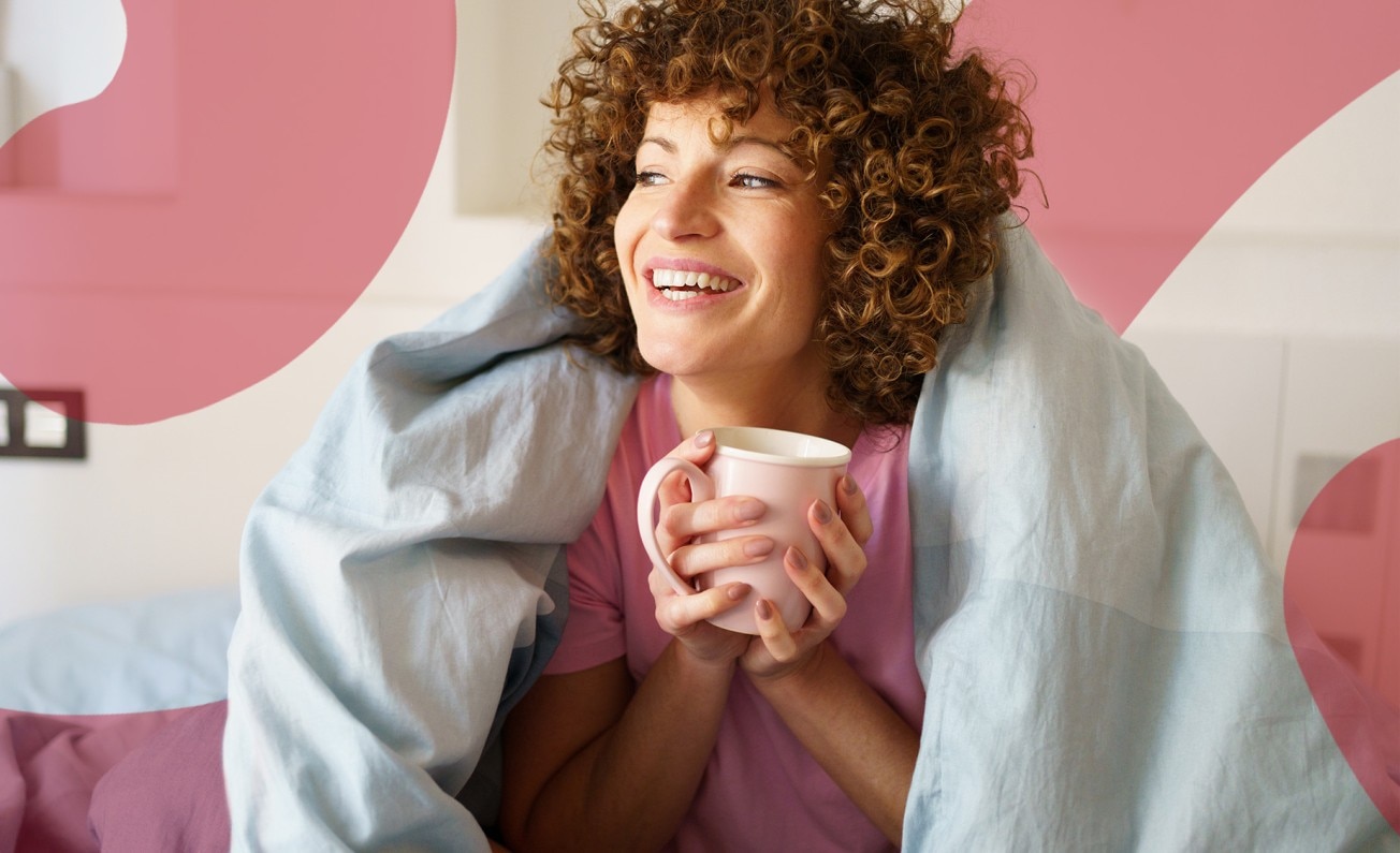 A smiling woman sits with a cup of hot drink and a duvet loosely around her shoulders