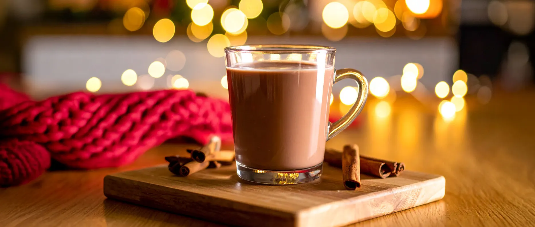 Glass mug of steaming Gingerbread Hot Chocolate made with KitKat, served on a wooden board with cinnamon sticks, festive ligh